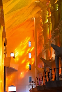 The image depicts an interior space with vibrant orange and yellow lighting illuminating the walls and architectural elements. The light casts complex shadows and patterns across the textured and angular surfaces. Tall vertical windows punctuate the walls, while silhouettes of railing structures add depth to the scene.