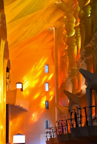 The image depicts an interior space with vibrant orange and yellow lighting illuminating the walls and architectural elements. The light casts complex shadows and patterns across the textured and angular surfaces. Tall vertical windows punctuate the walls, while silhouettes of railing structures add depth to the scene.