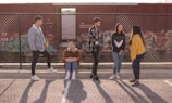 A group of five young individuals are interacting near a chain-link fence with a graffiti-covered train in the background. One person is sitting, while the others are standing, engaged in conversation and laughter. The background includes a red cargo train and some urban graffiti art.