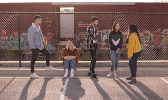 A group of five young individuals are interacting near a chain-link fence with a graffiti-covered train in the background. One person is sitting, while the others are standing, engaged in conversation and laughter. The background includes a red cargo train and some urban graffiti art.