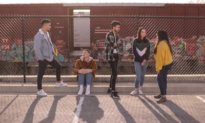 A group of five young individuals are interacting near a chain-link fence with a graffiti-covered train in the background. One person is sitting, while the others are standing, engaged in conversation and laughter. The background includes a red cargo train and some urban graffiti art.