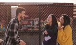 Three young adults are laughing and chatting in front of a chain-link fence with a colorful train in the background. They appear to be enjoying each other's company, with one wearing a plaid jacket, another in a dark green and purple sweater, and the third in a bright yellow sweater.
