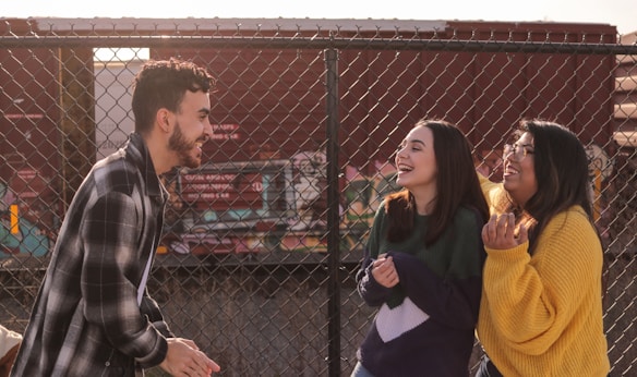 Three young adults are laughing and chatting in front of a chain-link fence with a colorful train in the background. They appear to be enjoying each other's company, with one wearing a plaid jacket, another in a dark green and purple sweater, and the third in a bright yellow sweater.