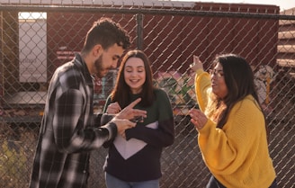 Three young adults are gathered in front of a chain-link fence, engaging in a lively conversation. The person on the left, wearing a black and white checkered shirt, is pointing with enthusiasm. The middle person, dressed in a green and purple sweater, is smiling broadly, conveying a sense of joy. The person on the right, in a bright yellow sweater, appears animated while gesturing with one hand.