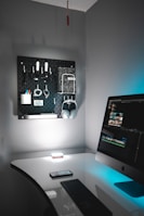 A tidy, modern home office setup featuring a sleek white desk with a black keyboard and a smartphone resting on it. An iMac is situated on the right, displaying a video editing software interface. On the wall, there is a black pegboard organizing various items including headphones, cables, and stationery. The lighting is soft with a blue accent, creating a calm and focused environment.
