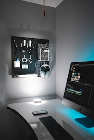 A tidy, modern home office setup featuring a sleek white desk with a black keyboard and a smartphone resting on it. An iMac is situated on the right, displaying a video editing software interface. On the wall, there is a black pegboard organizing various items including headphones, cables, and stationery. The lighting is soft with a blue accent, creating a calm and focused environment.