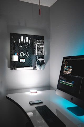A tidy, modern home office setup featuring a sleek white desk with a black keyboard and a smartphone resting on it. An iMac is situated on the right, displaying a video editing software interface. On the wall, there is a black pegboard organizing various items including headphones, cables, and stationery. The lighting is soft with a blue accent, creating a calm and focused environment.