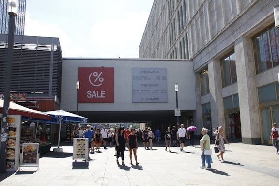 A bustling urban plaza with people walking in front of a large shopping center. A prominent sale sign with a percentage symbol is displayed on the building. Several individuals are casually dressed, and some are holding umbrellas for shade. There are outdoor cafe setups with awnings on the left.