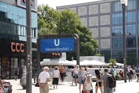 A busy urban square with people walking around and relaxing. A prominent blue sign for the U-Bahn indicates the Alexanderplatz station in a lively area surrounded by shops, including a CCC store. Trees and modern buildings with large windows line the background, adding to the cityscape.