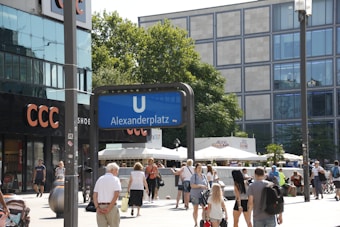 A busy urban square with people walking around and relaxing. A prominent blue sign for the U-Bahn indicates the Alexanderplatz station in a lively area surrounded by shops, including a CCC store. Trees and modern buildings with large windows line the background, adding to the cityscape.