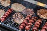 A grill loaded with several hamburger patties and skewered pieces of sausage. The meat is sizzling, and there is visible smoke rising from the hot grill. The patties appear juicy and slightly charred, while the sausage pieces are evenly spaced on wooden skewers.