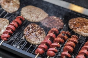 A grill loaded with several hamburger patties and skewered pieces of sausage. The meat is sizzling, and there is visible smoke rising from the hot grill. The patties appear juicy and slightly charred, while the sausage pieces are evenly spaced on wooden skewers.