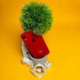 A red iPhone rests against a potted artificial plant with a green leafy top, surrounded by a small collection of accessories including a white USB charging cable, a charging adapter, and a pair of earphones. The background is a bright yellow, creating a vibrant and bold contrast.