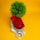 A red iPhone rests against a potted artificial plant with a green leafy top, surrounded by a small collection of accessories including a white USB charging cable, a charging adapter, and a pair of earphones. The background is a bright yellow, creating a vibrant and bold contrast.