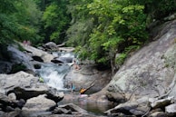A lush, green forest setting surrounds a cascading waterfall with clear pools of water. Several people are engaging in recreational activities like climbing rocks and wading in the water, suggesting a warm, leisurely day outdoors.