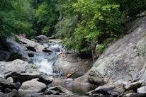 A lush, green forest setting surrounds a cascading waterfall with clear pools of water. Several people are engaging in recreational activities like climbing rocks and wading in the water, suggesting a warm, leisurely day outdoors.