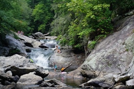 A lush, green forest setting surrounds a cascading waterfall with clear pools of water. Several people are engaging in recreational activities like climbing rocks and wading in the water, suggesting a warm, leisurely day outdoors.