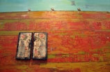 A textured painting with a vibrant, abstract background featuring shades of orange, red, and yellow alongside elements of blue and green. Two rectangular panels with floral and vase motifs occupy the lower left area. Small figures appear at the horizon, silhouetted against a vivid sky.