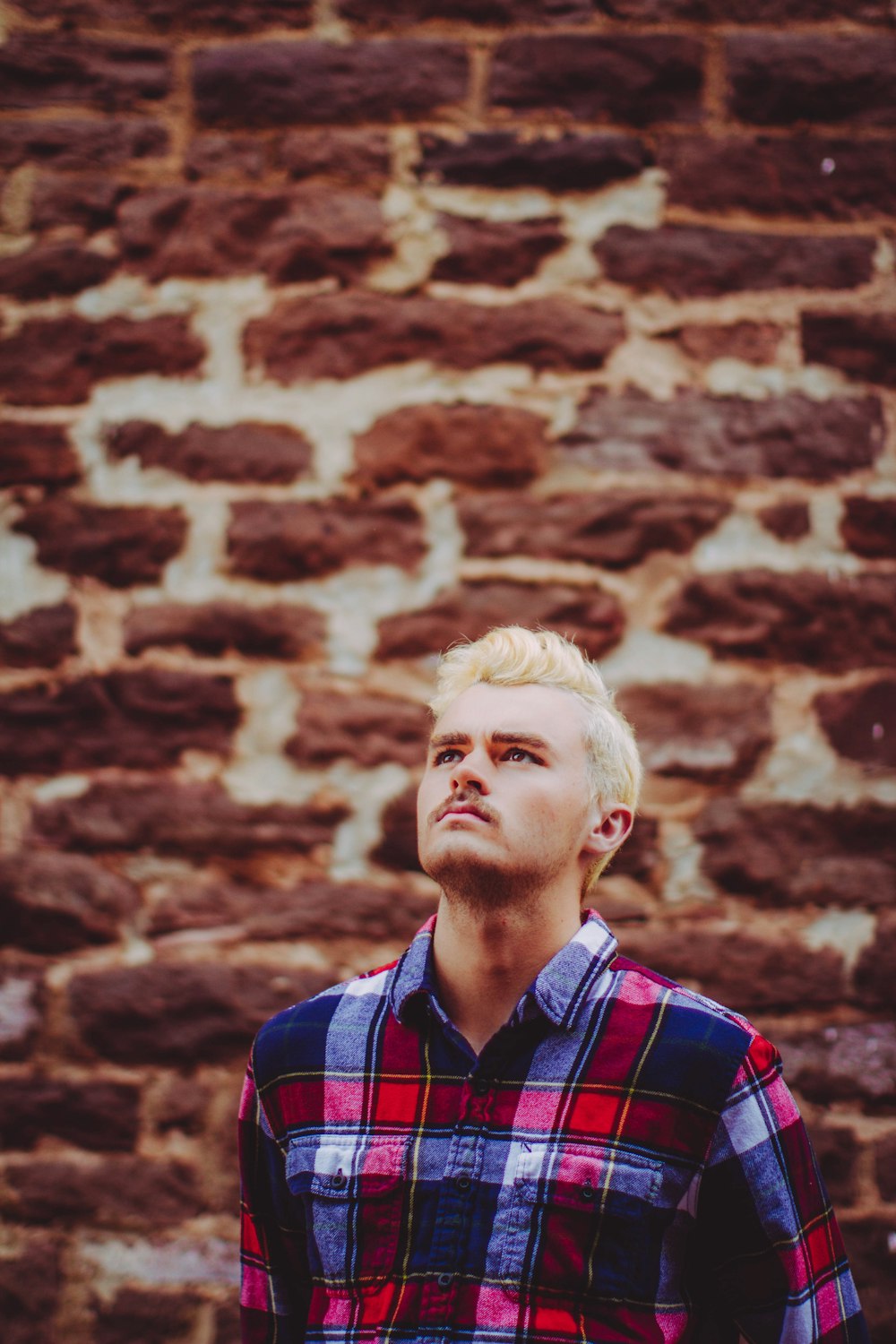 man in blue and red plaid shirt standing in front of brick wall