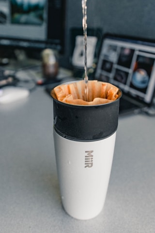 A stream of liquid is being poured into a coffee filter placed inside a travel mug. The mug has the brand name 'Miir' on it. The background shows a desk with two computer monitors and various items.