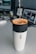A stream of liquid is being poured into a coffee filter placed inside a travel mug. The mug has the brand name 'Miir' on it. The background shows a desk with two computer monitors and various items.