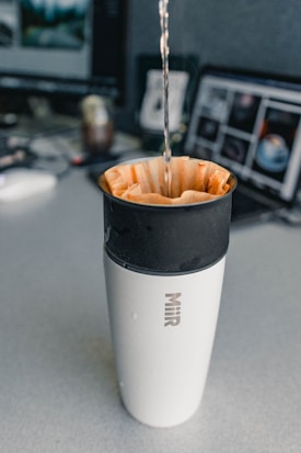 A stream of liquid is being poured into a coffee filter placed inside a travel mug. The mug has the brand name 'Miir' on it. The background shows a desk with two computer monitors and various items.