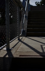A staircase with metal railings and a chain-link fence casting shadows on the ground. The light creates a contrast between the dark shadows and the illuminated parts of the stairs. The scene appears to be outdoors, with some greenery visible in the background.