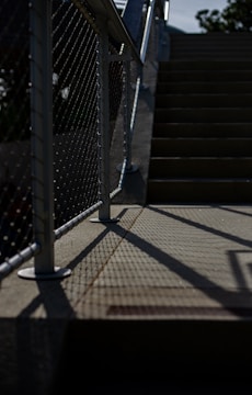 A staircase with metal railings and a chain-link fence casting shadows on the ground. The light creates a contrast between the dark shadows and the illuminated parts of the stairs. The scene appears to be outdoors, with some greenery visible in the background.