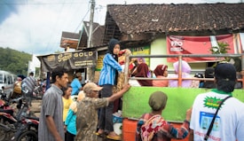 A group of people is gathered around an open truck, which is parked on the side of a street. Several people are standing on the truck, while others are interacting around it. There is a mix of men, women, and children dressed in casual clothing. The background features a rustic building with a tiled roof and signboards, along with other vehicles parked around.