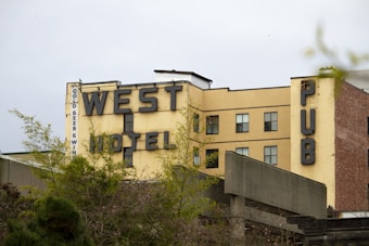 A multi-story building with large, bold lettering spelling 'WEST HOTEL' and 'PUB'. The building has a faded, yellow paint with sections of brick and multiple windows. Surrounding the building are some green trees and shrubs. The sky appears overcast.