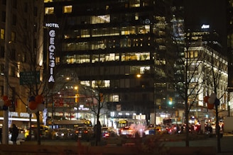 A bustling city scene at night with a prominent hotel sign lit up. Numerous office buildings with illuminated windows suggest activity inside. Streetlights and busy traffic fill the streets, while tree silhouettes and holiday-like decorations add to the urban ambiance.