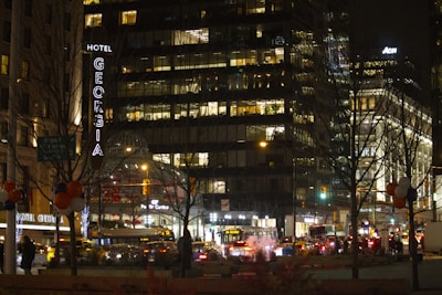 A bustling city scene at night with a prominent hotel sign lit up. Numerous office buildings with illuminated windows suggest activity inside. Streetlights and busy traffic fill the streets, while tree silhouettes and holiday-like decorations add to the urban ambiance.
