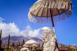 A stone statue with intricate carvings stands under a traditional Balinese parasol, with multiple temple structures and a mountainous landscape in the background. The sky is clear and bright blue, contrasting with the earthy tones of the statue and temples.