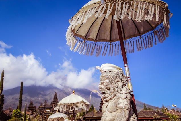 A stone statue with intricate carvings stands under a traditional Balinese parasol, with multiple temple structures and a mountainous landscape in the background. The sky is clear and bright blue, contrasting with the earthy tones of the statue and temples.