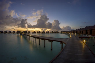 Brightly colored overwater bungalows reflecting softly on still lagoon waters at sunrise, framed by lush mangroves under a clear sky.