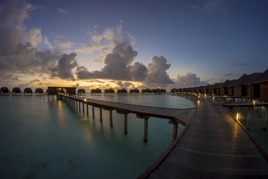 Brightly colored overwater bungalows reflecting softly on still lagoon waters at sunrise, framed by lush mangroves under a clear sky.