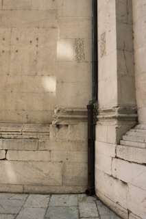 A section of an old stone wall with detailed carvings and an aged metal drainpipe running vertically along it. The stone is a light cream color, showing signs of wear and weathering. There is a pattern of small decorations carved into some stones, giving an ancient architectural feel.