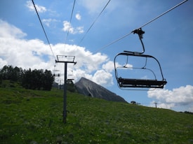 A chairlift system amid a green meadow with a mountainous backdrop. Empty chairlifts are suspended on cables, stretching towards a distant peak. The sky is blue with scattered clouds, and trees can be seen on the hillside.