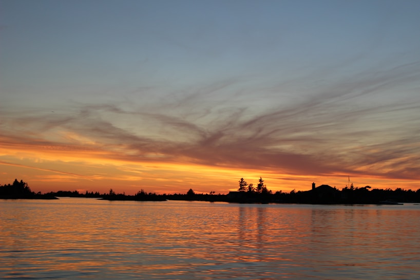 The image captures a serene sunset over a body of water, with the sky painted in shades of orange, yellow, and subtle blues. Silhouetted trees and a small structure are visible on the horizon. The water reflects the vibrant colors of the sky, enhancing the tranquil atmosphere.