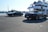 Two black luxury convertible cars are parked on a marina with a large yacht in the background. The setting is a bright and clear day, likely at a harbor with water visible in the distance. The area appears calm and upscale, suggesting a sense of affluence and leisure.