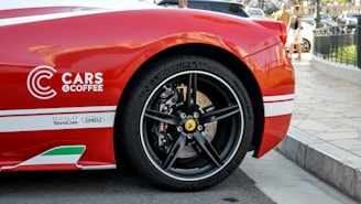 A close-up of the rear side of a red sports car with a focus on the wheel and the 'Cars & Coffee' logo. The car features sleek bodywork with white stripes and Italian flag accents. The background shows a paved street with several people and vehicles, indicating an event or gathering.