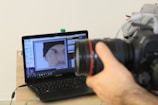 A hand is holding a DSLR camera focused on a laptop screen. The laptop screen displays an image editing software with a close-up photo of a person's face wearing a black beanie. The laptop is placed on a wooden table with a few items like a water bottle and some clothing in the background.