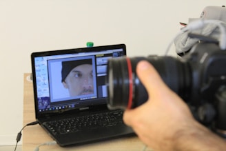 A hand is holding a DSLR camera focused on a laptop screen. The laptop screen displays an image editing software with a close-up photo of a person's face wearing a black beanie. The laptop is placed on a wooden table with a few items like a water bottle and some clothing in the background.