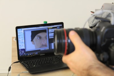 A hand is holding a DSLR camera focused on a laptop screen. The laptop screen displays an image editing software with a close-up photo of a person's face wearing a black beanie. The laptop is placed on a wooden table with a few items like a water bottle and some clothing in the background.