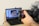 A hand is holding a DSLR camera focused on a laptop screen. The laptop screen displays an image editing software with a close-up photo of a person's face wearing a black beanie. The laptop is placed on a wooden table with a few items like a water bottle and some clothing in the background.