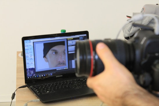 A hand is holding a DSLR camera focused on a laptop screen. The laptop screen displays an image editing software with a close-up photo of a person's face wearing a black beanie. The laptop is placed on a wooden table with a few items like a water bottle and some clothing in the background.