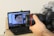 A hand is holding a DSLR camera focused on a laptop screen. The laptop screen displays an image editing software with a close-up photo of a person's face wearing a black beanie. The laptop is placed on a wooden table with a few items like a water bottle and some clothing in the background.