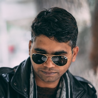 A man is wearing a black leather jacket and aviator sunglasses. His hair is short and styled, and he has a slight beard. The background is blurred, suggesting a shallow depth of field.