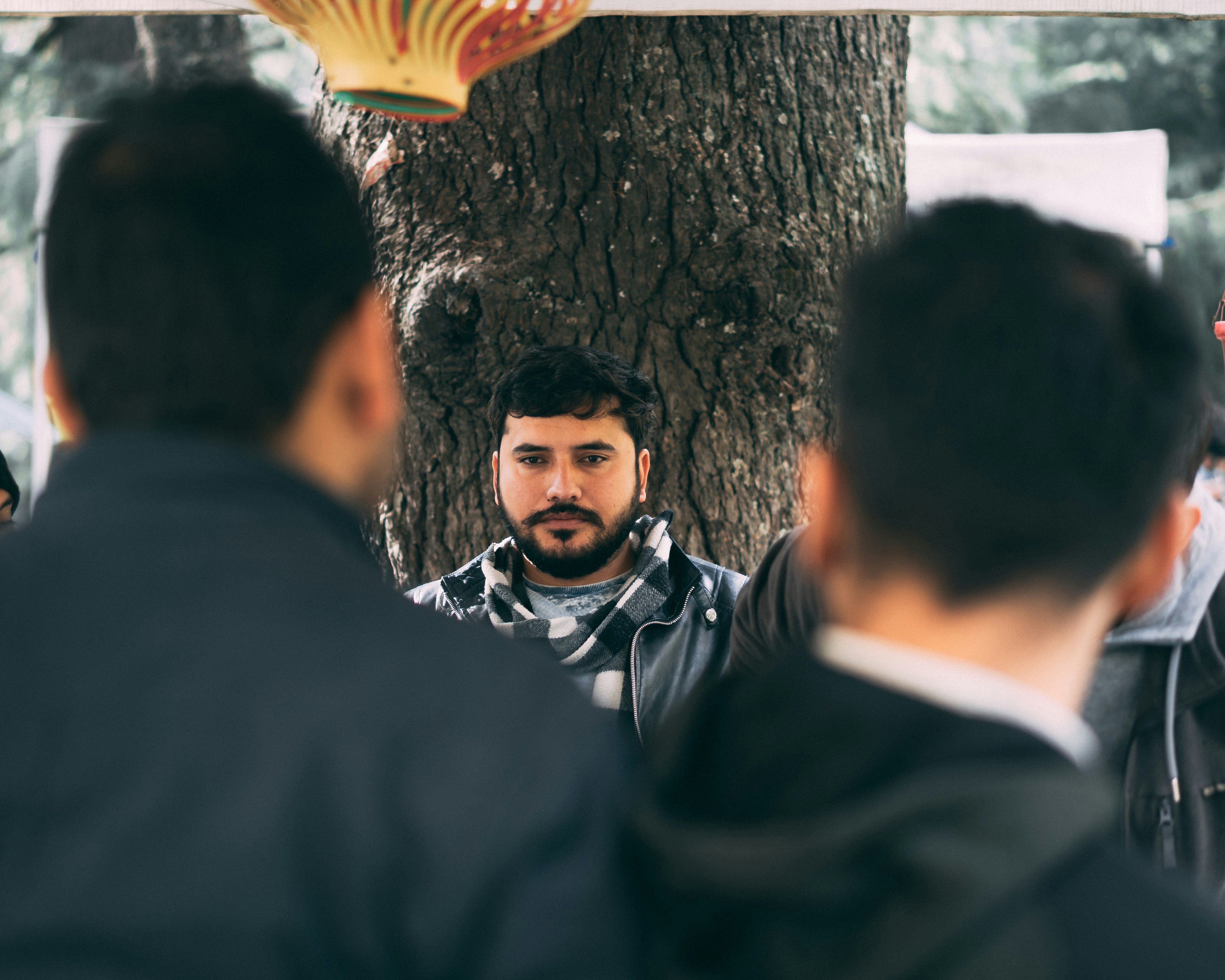 selective focus photography of man in gray and white plaid scarf leaning on tree during daytime