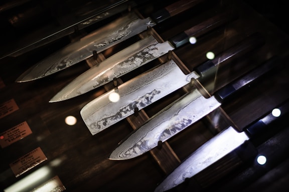 A set of Japanese chef knives with patterned blades is displayed in a wooden box. The blades show intricate patterns, possibly indicating Damascus steel. The knives have dark handles and are arranged neatly side by side. Reflections of lights can be seen on the glass covering.